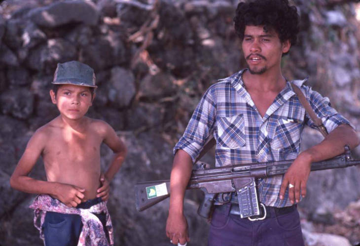 A guerrilla man and a child stand in front of a hillside,