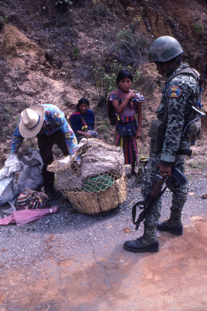 A soldier inspecting a Mayan man's basket in search of subversive materials