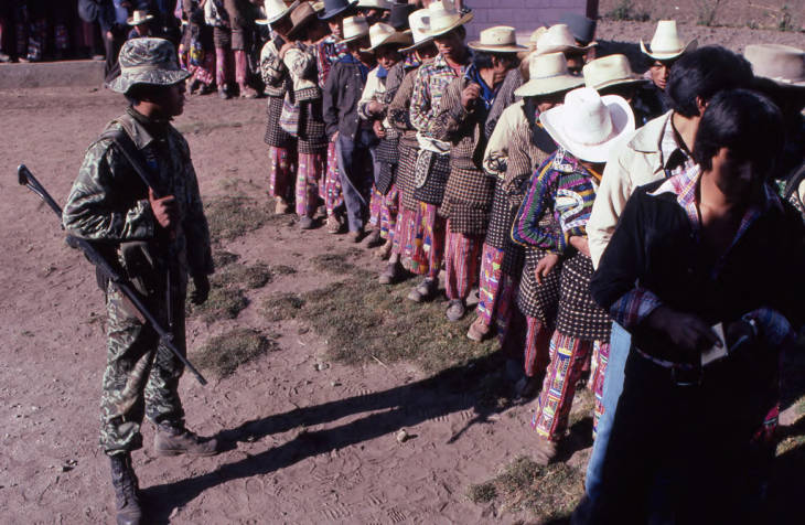 Mayan men in line to vote while a soldier watches them