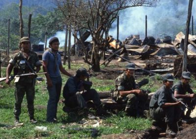 A group of men holding rifles are resting on wood barks. There is smoke in the background and a destroyed environment.